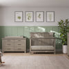 A serene nursery with light wood furniture, featuring a crib and a Child Craft F01792 Universal Dressing Kit in Crescent Grey on the dresser, green paneled walls, plant decor, nature prints, a rocking horse, and a large window for natural light.