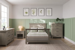A cozy bedroom with light green wainscoting, a wooden bed, nightstand, dresser, and the Child Craft Denman 4 Drawer Chest in Crescent Gray. Sunlight streams in, botanical prints hang above the bed, and a rug covers part of the wooden floor.