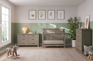 A bright, modern nursery features green and gray wooden furniture, including a crib and the Child Craft Denman Double Dresser in Crescent Gray. Nature-themed art prints adorn the walls, with a rocking horse on a beige rug near a large sunny window.