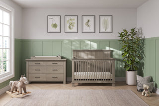 A nursery with light green and white walls, a wooden crib, rocking horse, potted plant, framed leaf art, teddy bear, a large window for natural light, and the Child Craft Denman Double Dresser in Crescent Gray with a changing pad.