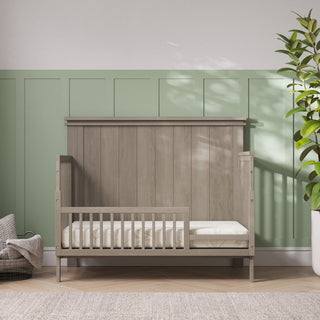 A modern nursery with a light wooden Child Craft crib featuring the F09503 - Toddler Guard Rail in Crescent Grey, set by a green paneled wall. Nearby are a wicker basket with a blanket and a tall leafy plant, bathed in sunlight.
