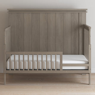 The Denman 4-in-1 Convertible Crib by Child Craft in Crescent Gray features a high slatted back, partial side rails, and includes a white mattress, set against a plain light wall on a light wood floor.