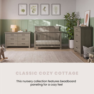 A cozy nursery with soft green beadboard paneling, a Child Craft Denman 4-in-1 Convertible Crib in Crescent Gray, dresser, chest, lamp, plush toys, and framed botanical art styled in a classic cottage theme.