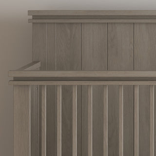 A close-up of the Child Craft Denman 4-in-1 Convertible Crib in Crescent Gray features vertical slats and a sturdy headboard, shown against a neutral wall.