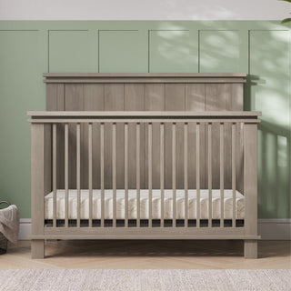 The Child Craft Denman 4-in-1 Convertible Crib in Crescent Gray, with vertical slats and a high back panel, stands against a green paneled wall. A light mattress is inside, and a basket rests on the floor nearby amid soft natural light.