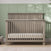 The Child Craft Denman 4-in-1 Convertible Crib in Crescent Gray, with vertical slats and a high back panel, stands against a green paneled wall. A light mattress is inside, and a basket rests on the floor nearby amid soft natural light.