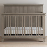 The Child Craft Denman 4-in-1 Convertible Crib in Crescent Gray features vertical slats and a solid headboard, set on a light wood floor against a white wall. The crib includes a white mattress inside.