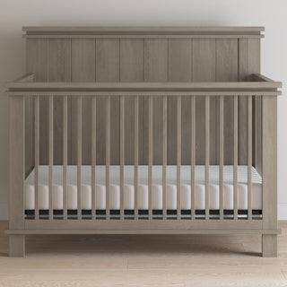 The Child Craft Denman 4-in-1 Convertible Crib in Crescent Gray features vertical slats and a solid headboard, set on a light wood floor against a white wall. The crib includes a white mattress inside.