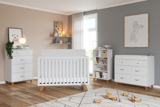 A modern nursery with white furniture, including the Child Craft Soho 4 Drawer Chest in White/Natural. Wooden toys rest on a geometric rug, animal art decorates beige and white walls, and sunlight pours through the window.
