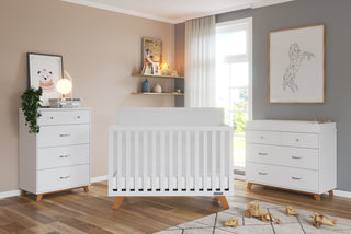 A modern nursery features a white crib, Child Craft Soho 4 Drawer Chests in white/natural on each side, wooden floors, neutral walls, a window with sheer curtains, decor on shelves, and toys scattered on the floor.