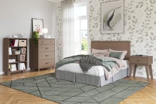 A modern bedroom features a bed, dresser, and the Soho Nightstand in Dusty Heather by Child Craft beneath leafy wallpaper. Sunlight streams in through a big window, and a green geometric rug brightens the wood floor.