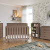 A modern nursery with light wood furniture, including a crib, two dressers—one topped with the Child Craft F01792 Universal Changing Table Topper in Dusty Heather—and a bookshelf. Toys cover a green rug, while leafy wallpaper and sunlight brighten the space.