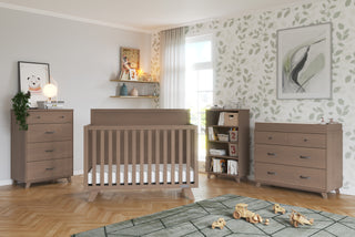 A modern nursery with light wood furniture, including a crib, two dressers—one topped with the Child Craft F01792 Universal Changing Table Topper in Dusty Heather—and a bookshelf. Toys cover a green rug, while leafy wallpaper and sunlight brighten the space.