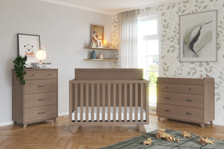 A modern nursery features wood furniture: a crib, two dressers—one topped with the Child Craft F01792 Universal Changing Table Topper in Dusty Heather—and shelves. Toys are on the floor, sunlight streams through sheer curtains, and leafy wallpaper covers a wall.