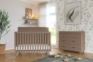A cozy nursery featuring a wooden crib, matching dresser topped with the Child Craft F01792 Universal Changing Table Topper in Dusty Heather, leafy wallpaper, a green rug with wooden toys, shelves with books and decor, a large plant, and sunlight through the window.