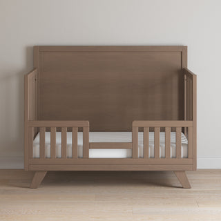 The Child Craft Toddler Guard Rail in Dusty Heather, designed with baby-safe finishes and side guardrails, is shown with an open center space, placed against a plain white wall on a wooden floor.