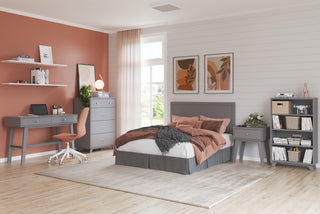 A cozy bedroom features a gray bed, rust-colored bedding, a gray desk and chair, bookshelves, and modern decor. Light wood floors, white walls, and a large window brighten the space. The Child Craft Soho 4-in-1 Crib in Cool Gray adds versatility.