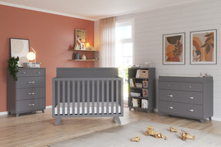 A modern nursery featuring the Child Craft Soho 4-in-1 Flat Top Convertible Crib in Cool Gray, paired with a dresser, changing table, bookshelf, and toys on a light wood floor. White and terracotta walls with art and sheer curtains create warmth.