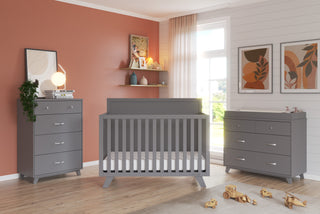 A modern nursery featuring the Child Craft Soho 4-in-1 Flat Top Convertible Crib in Cool Gray, a matching dresser and changing table, with orange and white walls, shelves, framed art, toys, a plant, and sunlight streaming through sheer curtains.