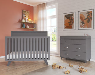A modern nursery with the Child Craft Soho 4-in-1 Flat Top Convertible Crib in Cool Gray, matching dresser, wooden toys, art prints, shelves with books and decor, white walls, sheer curtains, and a rust-colored accent wall for a cozy feel.