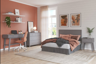 A modern bedroom with a gray bed, rust bedding, matching desk and chair, dresser, nightstand, wall art, floating shelves, plants, large window with sheer curtains, and the Child Craft Soho 4-in-1 Flat Top Convertible Crib in Cool Gray for a modern nursery touch.