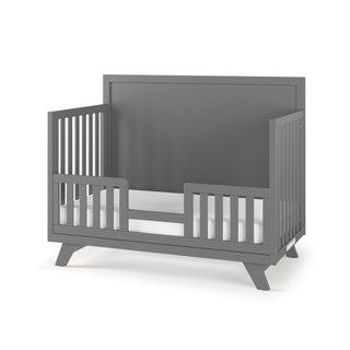 The Child Craft Soho 4-in-1 Flat Top Convertible Crib in Cool Gray features mid-century modern design, open side slats, a partial front safety rail, an elevated headboard, and angled legs—perfect for a stylish nursery.