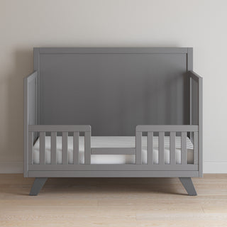 The Child Craft Soho 4-in-1 Flat Top Convertible Crib in Cool Gray stands stylishly against a light-colored wall on a light wooden floor, featuring open front and side railings for a modern nursery look.