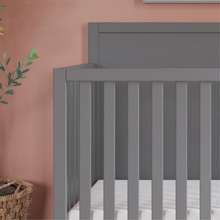 The Child Craft Soho 4-in-1 Flat Top Convertible Crib in Cool Gray stands against a pink wall, with a woven basket and a leafy plant to the left. A white mattress rests on the crib's base, showcasing its mid-century modern design with vertical slats.