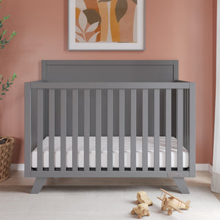 The Child Craft Soho 4-in-1 Flat Top Convertible Crib in Cool Gray with a white mattress sits against a muted pink wall. Abstract art hangs above, a plant basket is on the left, and wooden toys like a plane are scattered on the floor in front.