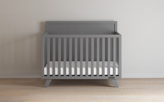 The Child Craft Soho 4-in-1 Flat Top Convertible Crib in Cool Gray, featuring vertical slats and a white mattress, stands on a wooden floor against a light wall, adding modern versatility and style to your nursery.