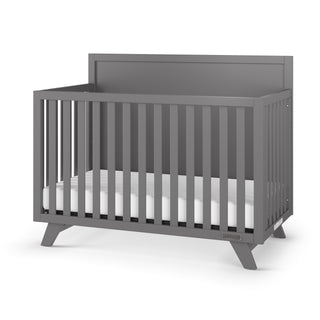 The Child Craft Soho 4-in-1 Flat Top Convertible Crib in Cool Gray features vertical slats, a solid back panel, and angled legs for a modern look. Shown with a white mattress, it's an ideal centerpiece for any nursery.