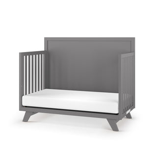 The Child Craft Soho 4-in-1 Flat Top Convertible Crib in Cool Gray features open slatted sides, a solid high back panel, angled legs, and a white mattress for a minimalist, mid-century modern look in your nursery.