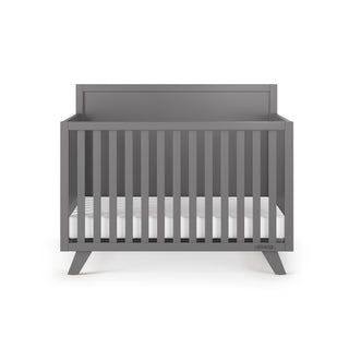 The Child Craft Soho 4-in-1 Flat Top Convertible Crib in Cool Gray features vertical slats, a high back panel, and angled legs, shown with a white mattress—stylish and versatile nursery furniture for any modern space.
