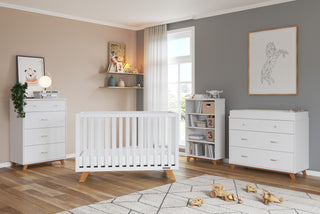 A modern nursery features the Child Craft Soho 4 Drawer Chest in white/natural. White furniture, a plush rug, and geometric animal prints complete the space, with light streaming through a large window onto wooden floors.