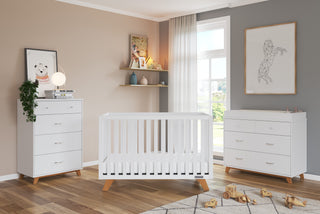 A modern nursery featuring a Child Craft Soho 4 Drawer Chest in white/natural, a white crib at the center, toys on a wood floor, decorated shelves, wall art, and sunlight filtering through sheer curtains.