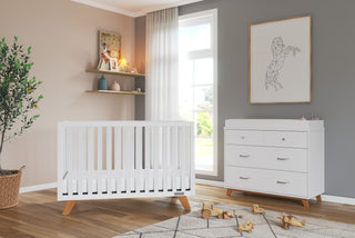 A modern nursery features the Child Craft Soho 4-in-1 Convertible Crib in White/Natural, a matching dresser, framed geometric horse art, wooden toys on a white rug, a potted plant, shelves with books and decor, and large windows with natural light.