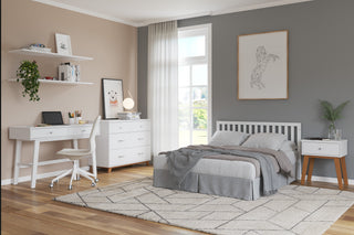 A modern bedroom features the Child Craft Soho 4-in-1 Convertible Crib in White/Natural, gray accent wall, white furniture, gray bedding, desk with chair, shelves with books/plants, dresser, geometric art above the bed, and sunlight from a window.