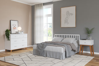 A modern bedroom with gray and beige decor features a neatly made bed, geometric rug, large windows, minimalist art, and a Child Craft Soho 3-Drawer Dresser in White/Natural topped with books and a lamp, plus a plant on the nightstand.
