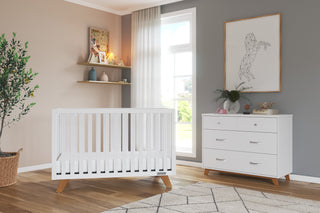 A modern nursery featuring the Child Craft Soho 4-in-1 Convertible Crib in White/Natural, a white dresser with wooden legs, a plant in a basket, sheer window curtains, and a wall shelf with toys and decor above the crib. Geometric horse art hangs above the dresser.
