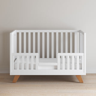 The Child Craft Soho 4-in-1 Convertible Crib in White/Natural features wooden legs, open side slats, and partial front guard rails, set against a light wall and floor for a modern toddler bed look.