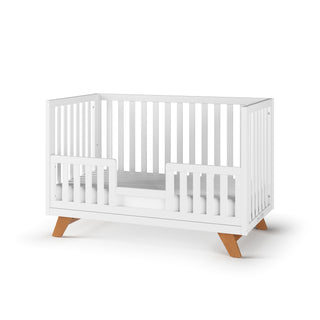 The Child Craft Soho 4-in-1 Convertible Crib in White/Natural features slatted sides, an open front panel with a low safety rail, and wooden legs. Displayed on a white background.