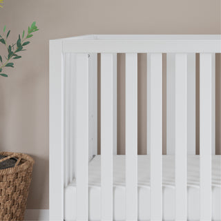 The Child Craft Soho 4-in-1 Convertible Crib in White/Natural is set against a beige wall. To the left, a woven basket and a green leafy plant are partially visible. The crib features vertical slats and a plain white mattress.
