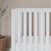 The Child Craft Soho 4-in-1 Convertible Crib in White/Natural is set against a beige wall. To the left, a woven basket and a green leafy plant are partially visible. The crib features vertical slats and a plain white mattress.