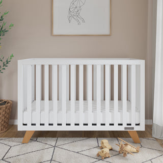 The Child Craft Soho 4-in-1 Convertible Crib in White/Natural features wooden legs and stands on a geometric rug in a softly lit nursery, with wooden toys, a plant, and a basket nearby, and framed art above on the beige wall.