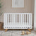 The Child Craft Soho 4-in-1 Convertible Crib in White/Natural features wooden legs and stands on a geometric rug in a softly lit nursery, with wooden toys, a plant, and a basket nearby, and framed art above on the beige wall.