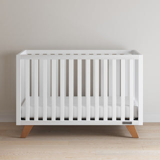 The Child Craft Soho 4-in-1 Convertible Crib in White/Natural features a minimalist design with horizontal slats and wooden legs, standing against a light wall on a light wood floor.