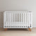 The Child Craft Soho 4-in-1 Convertible Crib in White/Natural features a minimalist design with horizontal slats and wooden legs, standing against a light wall on a light wood floor.