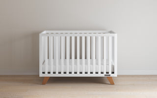 The Child Craft Soho 4-in-1 Convertible Crib in White/Natural features minimalist white design, wooden legs, and vertical slats. It stands on a light wood floor against a plain wall, featuring a simple white mattress.