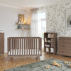 A modern nursery with light wood furniture: a crib, two dressers—one topped with the Child Craft Universal Changing Table Topper in Dusty Heather—a bookshelf of toys, playful wall art, leaf-patterned wallpaper, and a sunlit window.