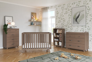 A modern nursery with light wood furniture: a crib, two dressers—one topped with the Child Craft Universal Changing Table Topper in Dusty Heather—a bookshelf of toys, playful wall art, leaf-patterned wallpaper, and a sunlit window.
